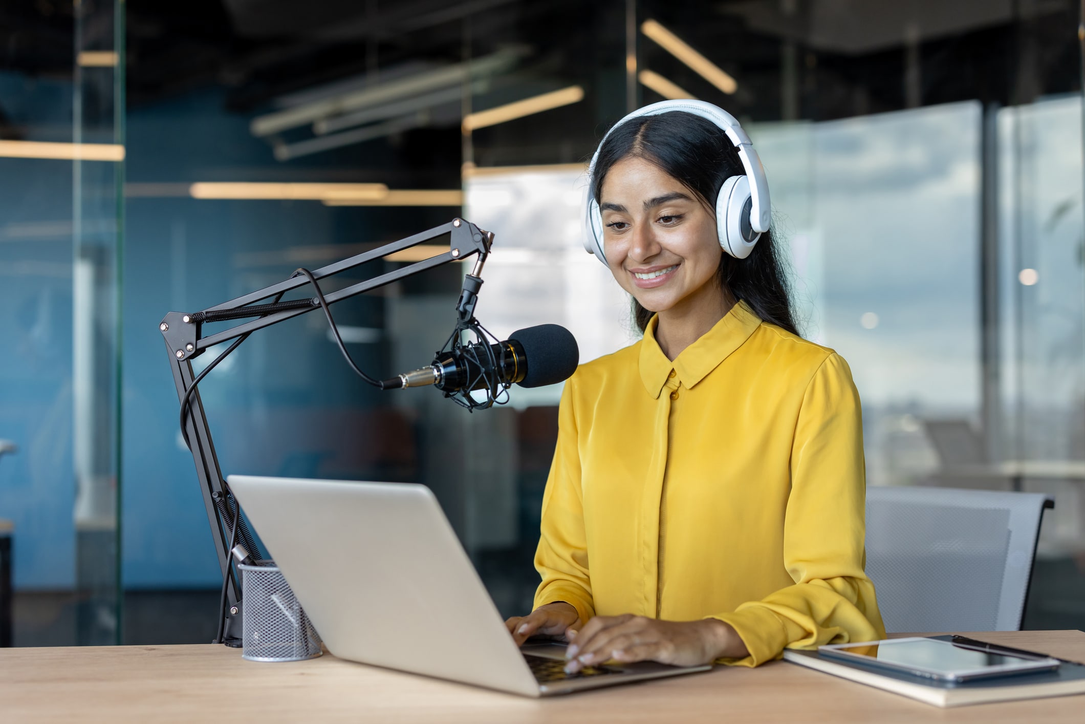 Smiling Indian woman sitting at a desk in the office wearing headphones, working on a laptop and talking into a microphone